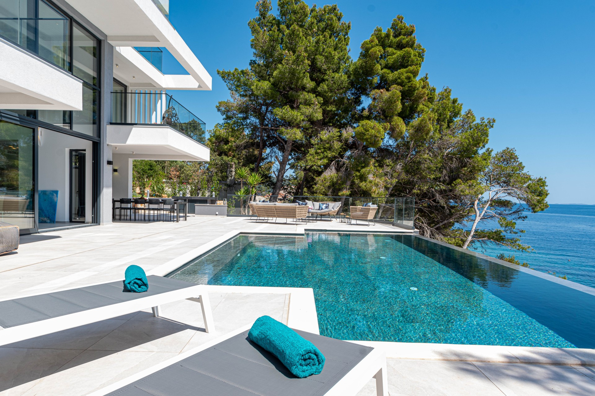 Modern luxury Villa Sansarea with a private swimming pool and spacious sun terrace, featuring outdoor lounge and dining areas, surrounded by pine trees and overlooking the crystal-clear sea.