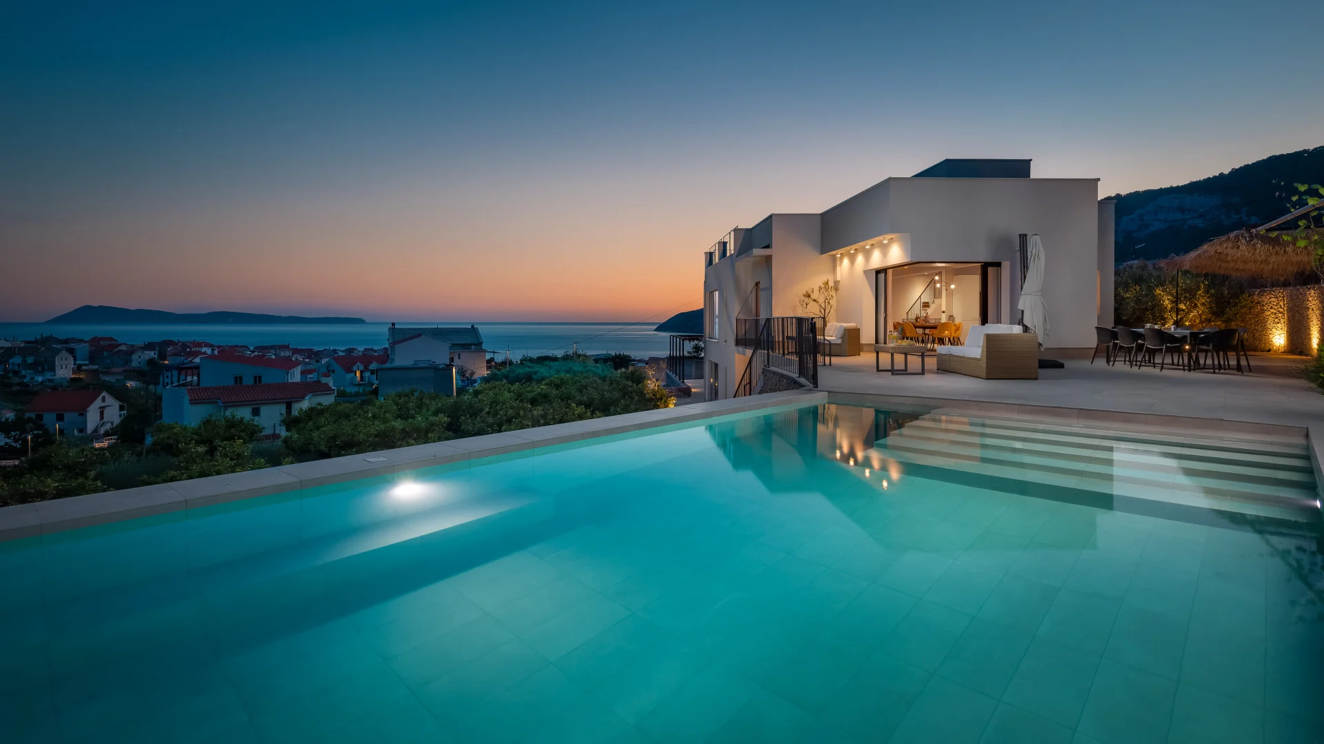 Modern luxury Villa Envivo with an infinity pool at sunset, featuring a spacious terrace with outdoor dining and lounge area, overlooking the sea and coastal village, with warm ambient lighting and panoramic views.