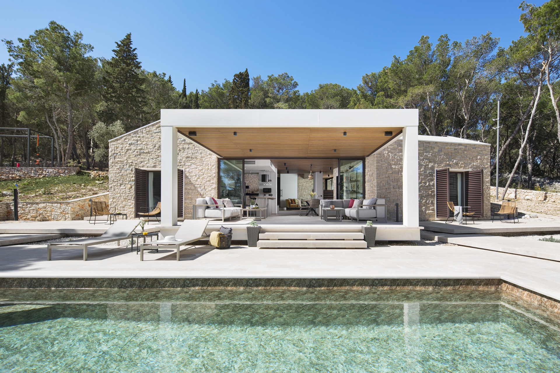 Villa en pierre de luxe au design moderne avec piscine, terrasse couverte dotée d'un salon extérieur, entourée de pins.