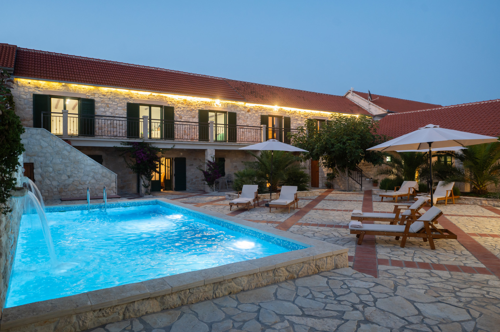 Domaine en pierre dalmate traditionnel avec piscine illuminée et cascade, vaste cour et solarium au crépuscule.