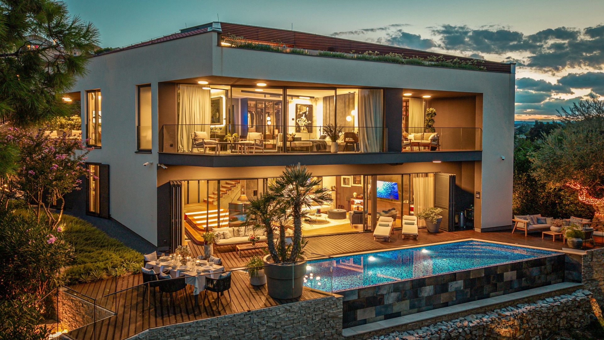 Villa di lusso ultramoderna su più piani con piscina illuminata, pareti in vetro e zona pranzo esterna al crepuscolo.