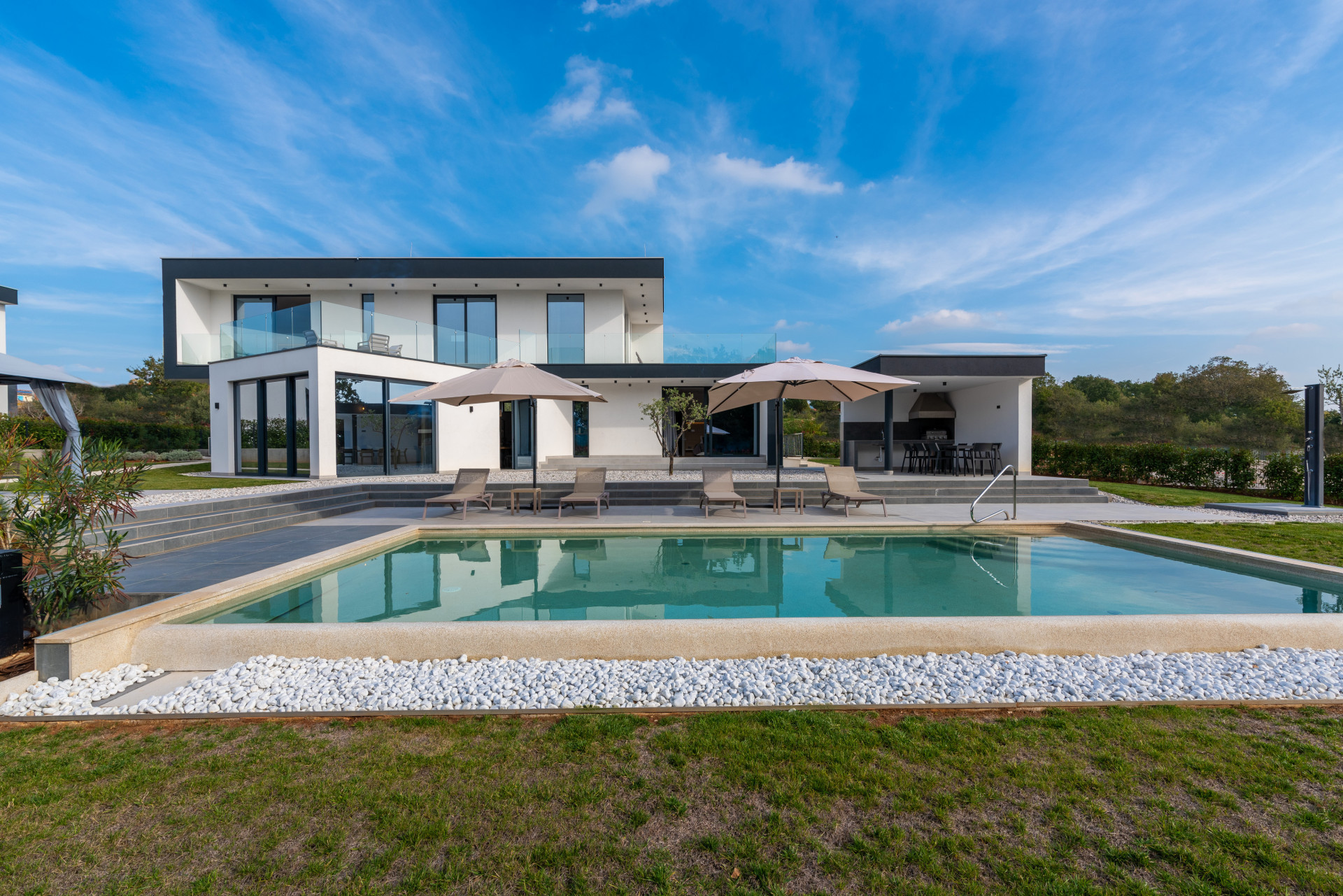 Modern two-story luxury villa with an outdoor swimming pool, sun deck, white pebbles, and an outdoor summer kitchen under a clear sky.