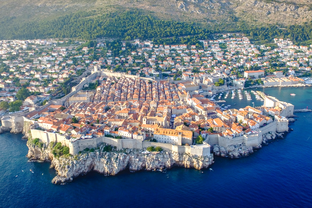 Veduta panoramica aerea della Città Vecchia di Dubrovnik circondata da mura medievali in pietra e dal profondo Mar Adriatico blu, con tetti in terracotta e colline verdi sullo sfondo.