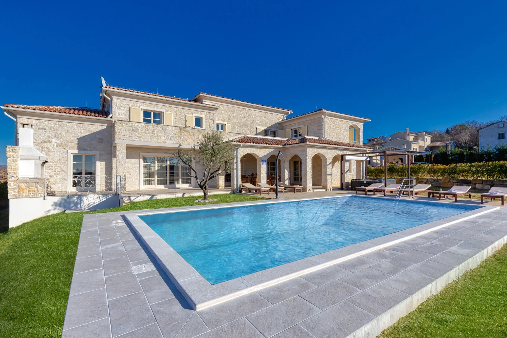 Villa di pietra di lusso in Istria con una grande piscina esterna, ampia terrazza prendisole con lettini, patio coperto e giardino paesaggistico sotto un cielo azzurro sereno.