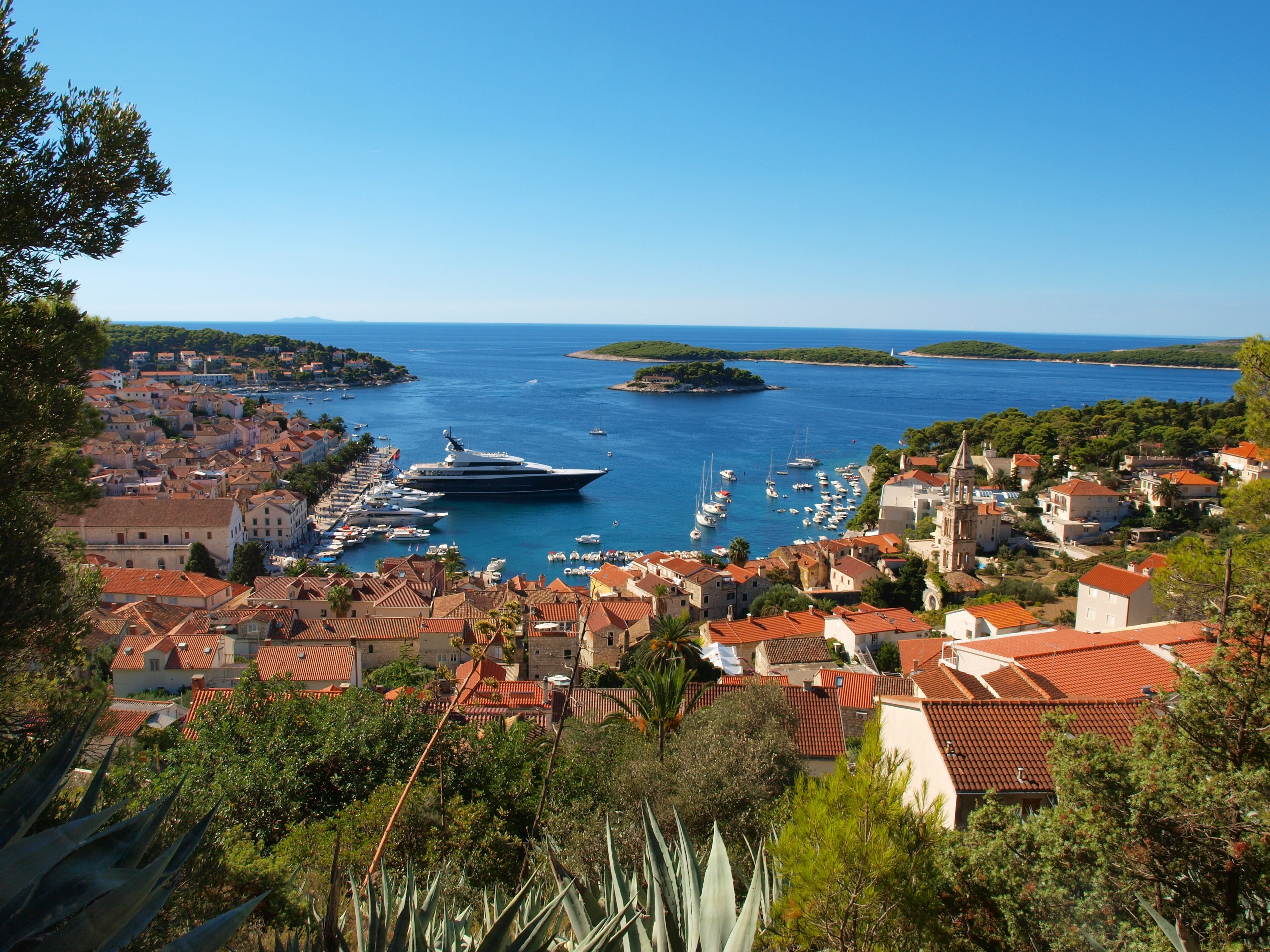 Veduta panoramica della città di Hvar con tetti in terracotta, un vivace porto turistico con yacht e barche, il mare Adriatico cristallino e isole verdi sotto un cielo azzurro intenso.