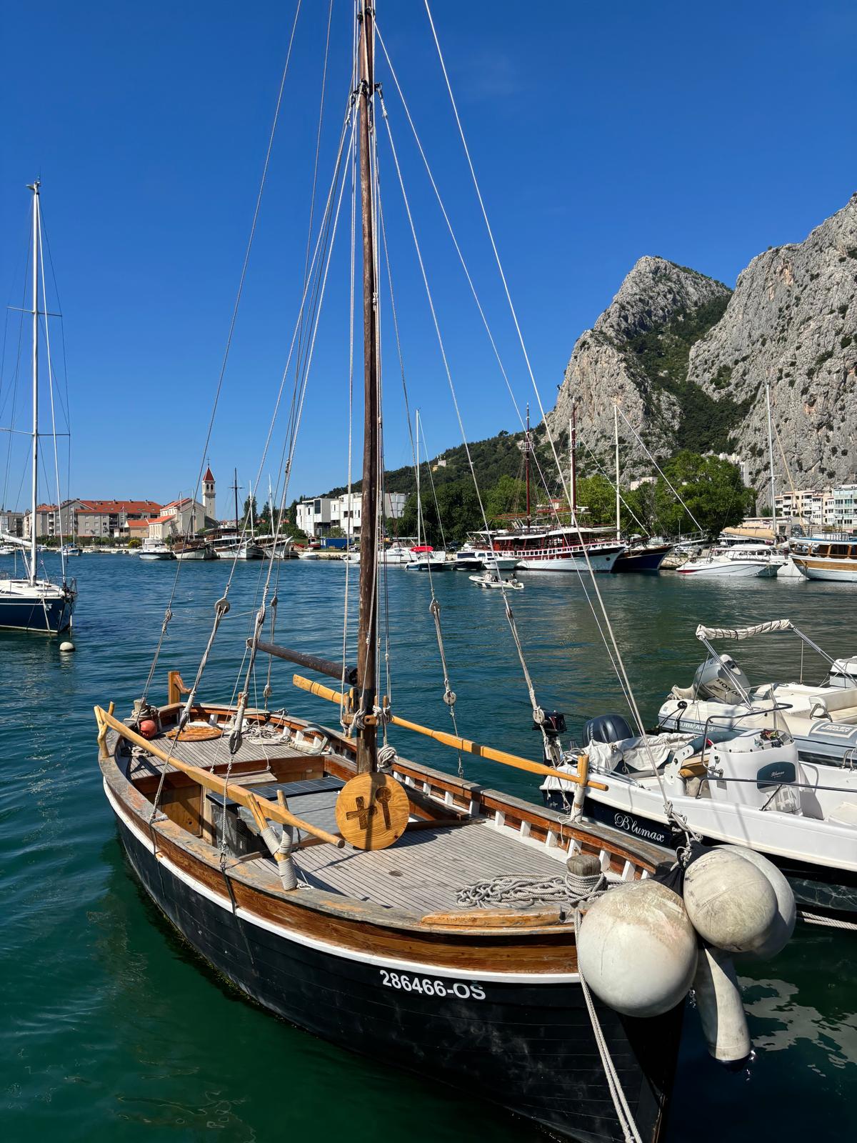 Traditional wooden sailboat moored in Omiš harbor, surrounded by yachts and boats with dramatic rocky mountains rising in the background.