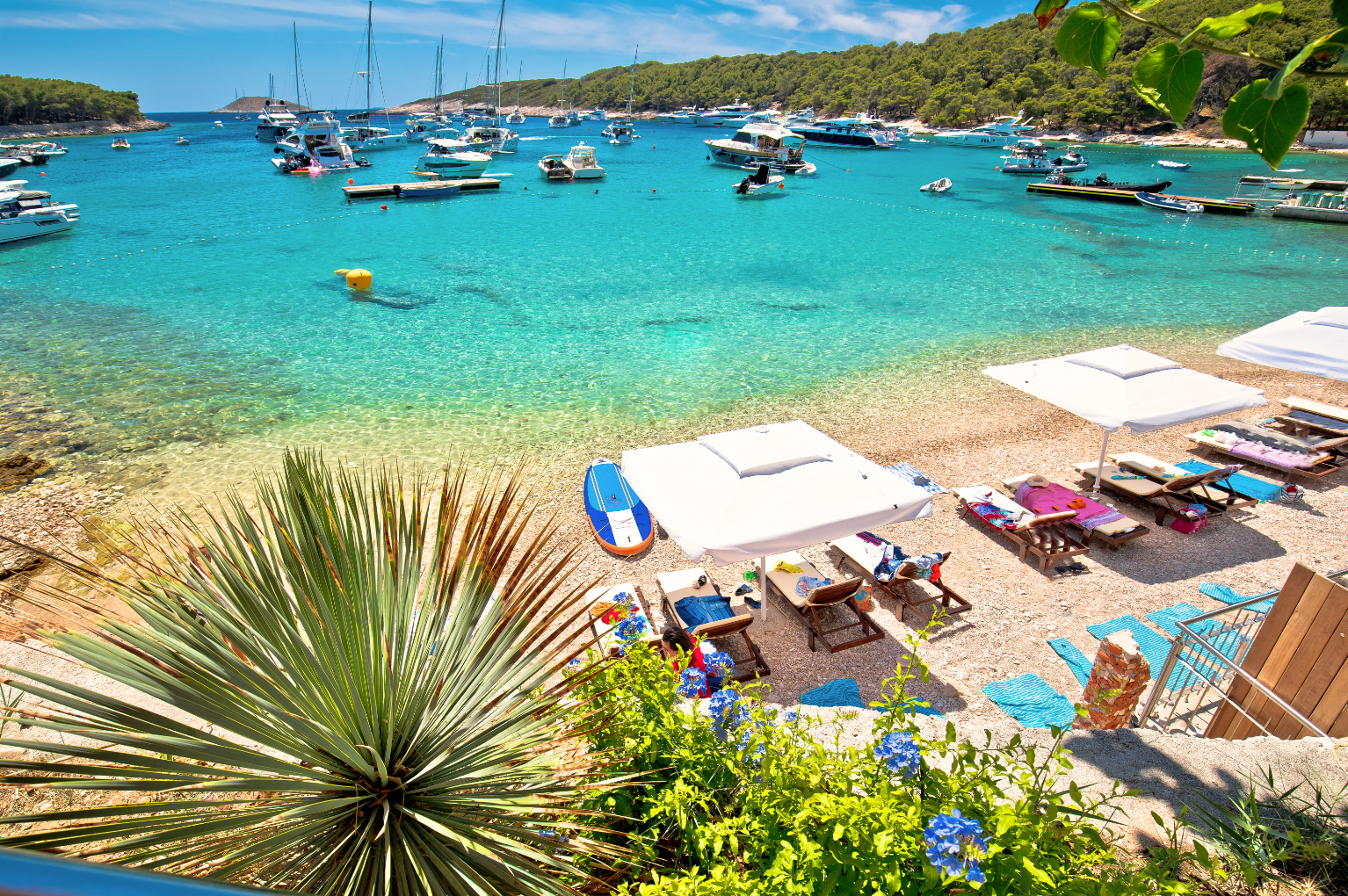 Palmižana Strand auf den Pakleni-Inseln bei Hvar, mit kristallklarem türkisfarbenem Wasser, Liegen mit weißen Sonnenschirmen und Yachten, die in einer malerischen mediterranen Bucht vor Anker liegen.