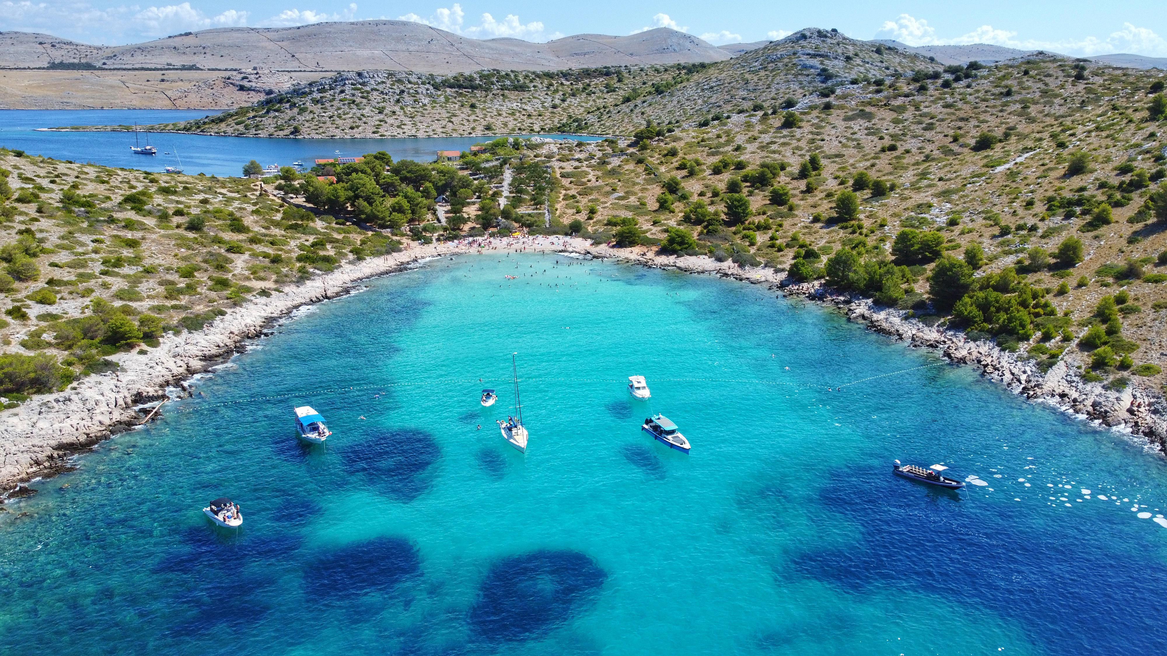 Aerial view of Lojena Bay on Levrnaka Island in Kornati National Park, featuring crystal-clear turquoise water, anchored boats, and a white pebble beach surrounded by a rugged Mediterranean landscape.