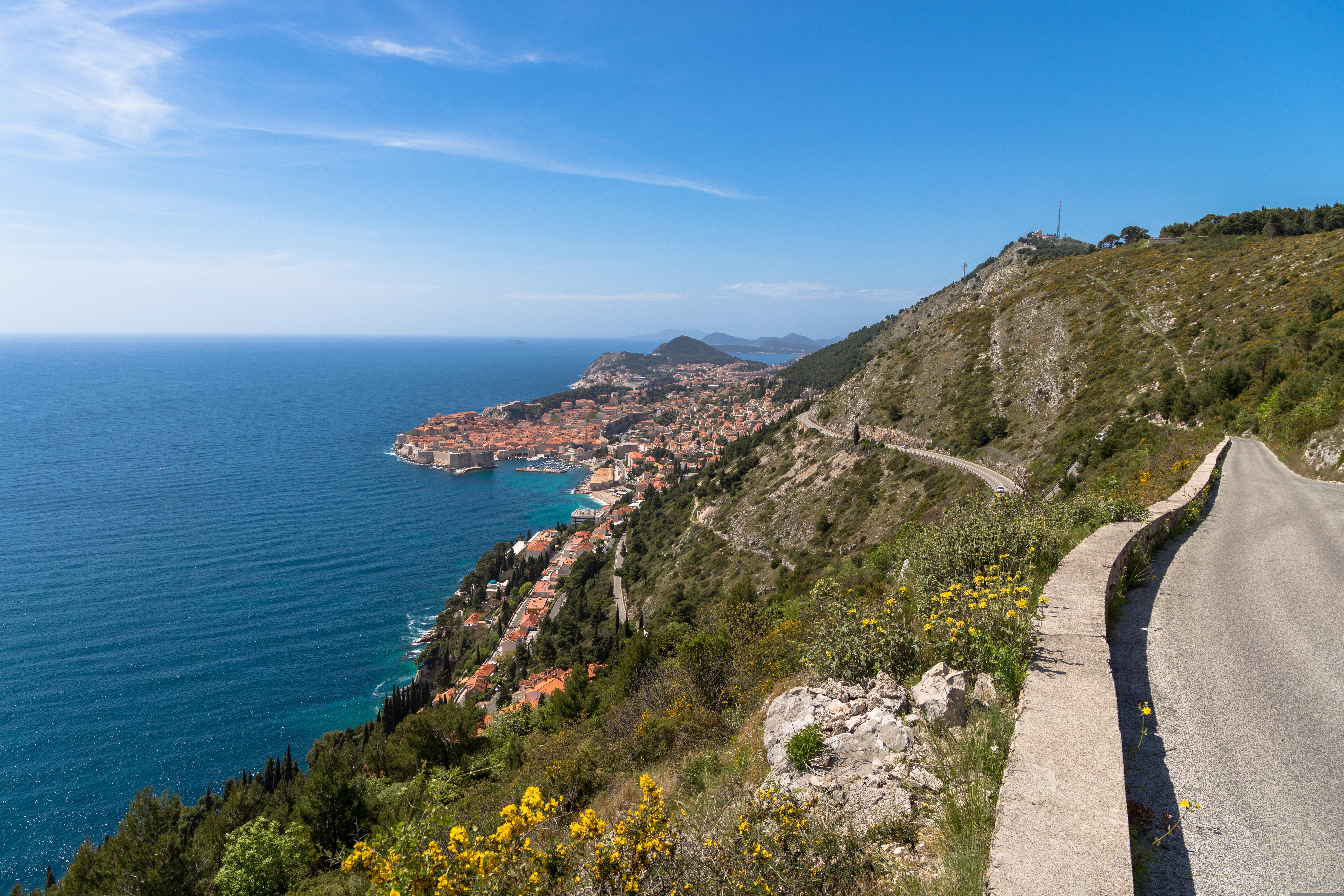 Scenic coastal road overlooking Dubrovnik and the Adriatic Sea, surrounded by rugged hills and Mediterranean landscape under a clear blue sky.