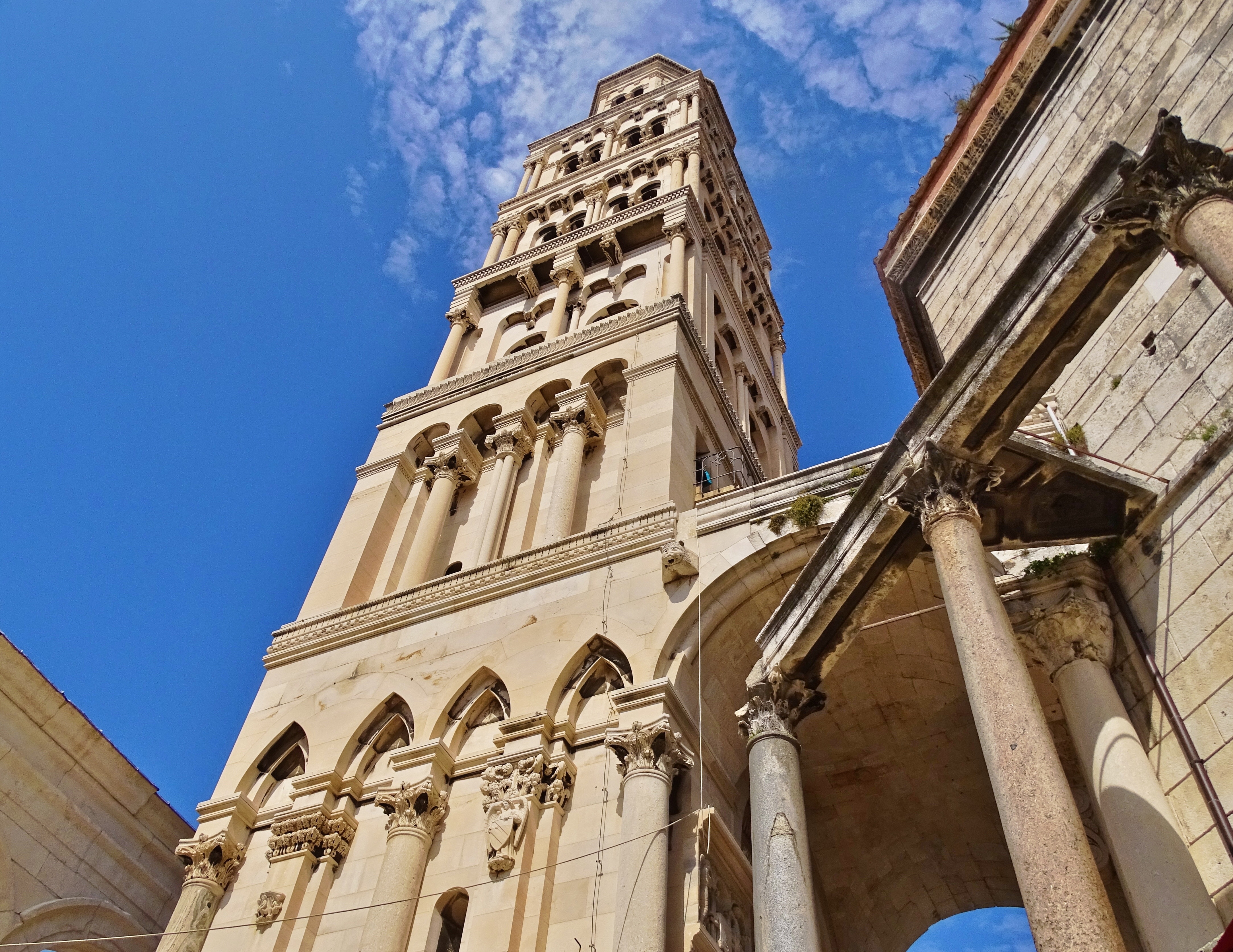 Low-angle view of the historic bell tower of Diocletian’s Palace in Split, Croatia, framed by ancient stone columns against a bright blue sky.