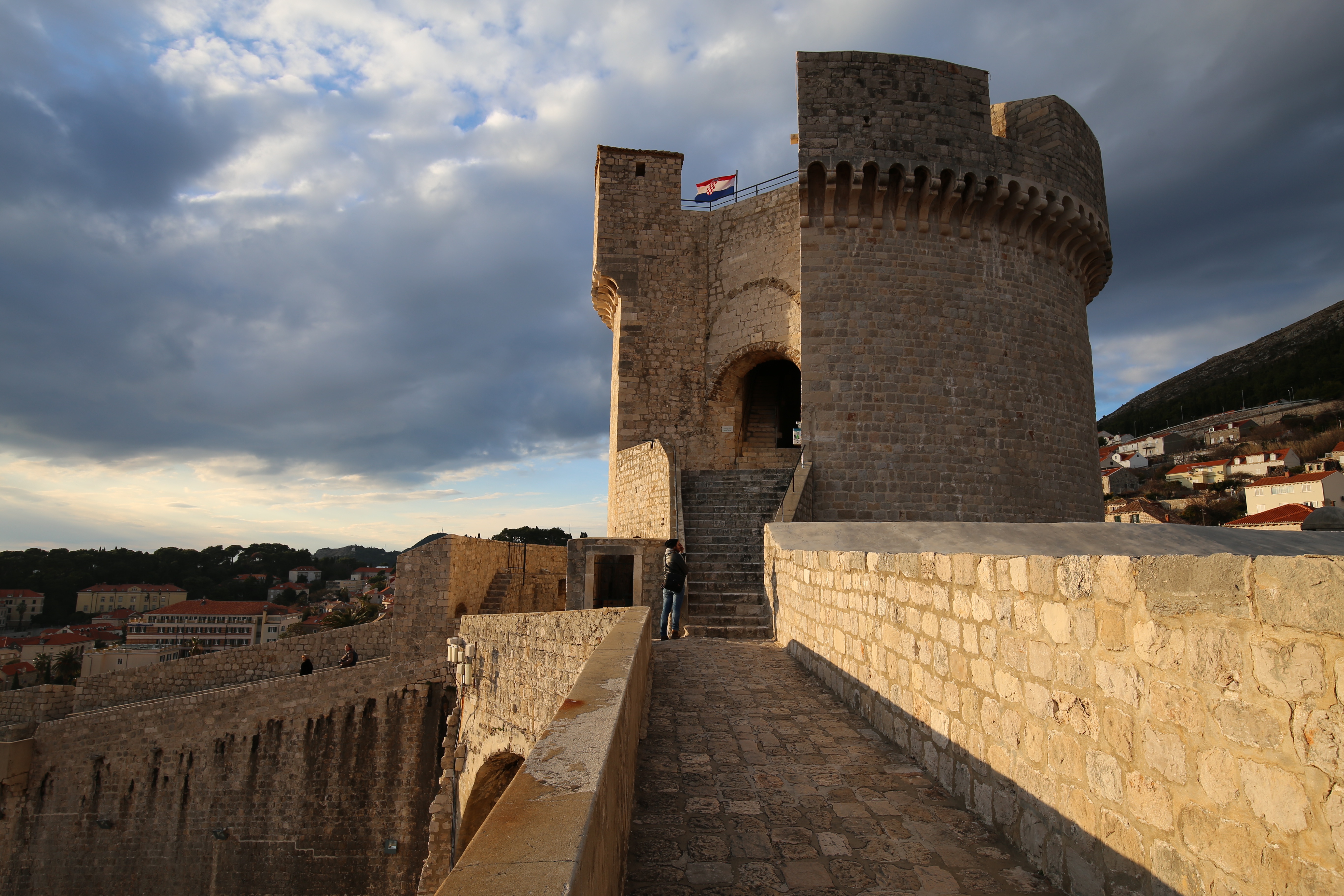Historic stone tower and fortified city walls of Dubrovnik, Croatia, illuminated by warm sunlight beneath a dramatic cloudy sky.