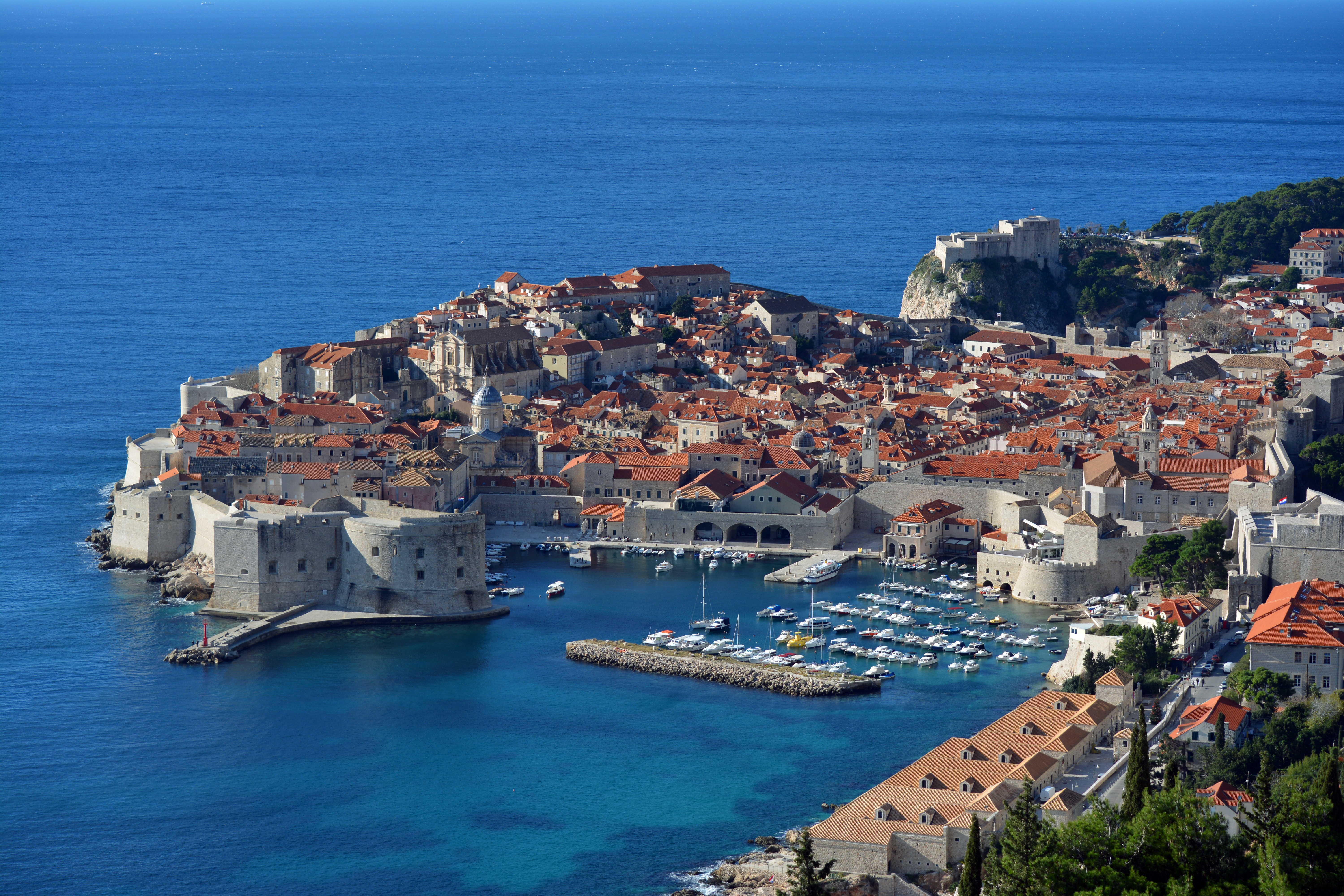 Aerial panoramic view of Dubrovnik Old Town, a UNESCO World Heritage Site, featuring medieval stone walls, the historic harbor, red-tiled rooftops, and the Adriatic Sea.