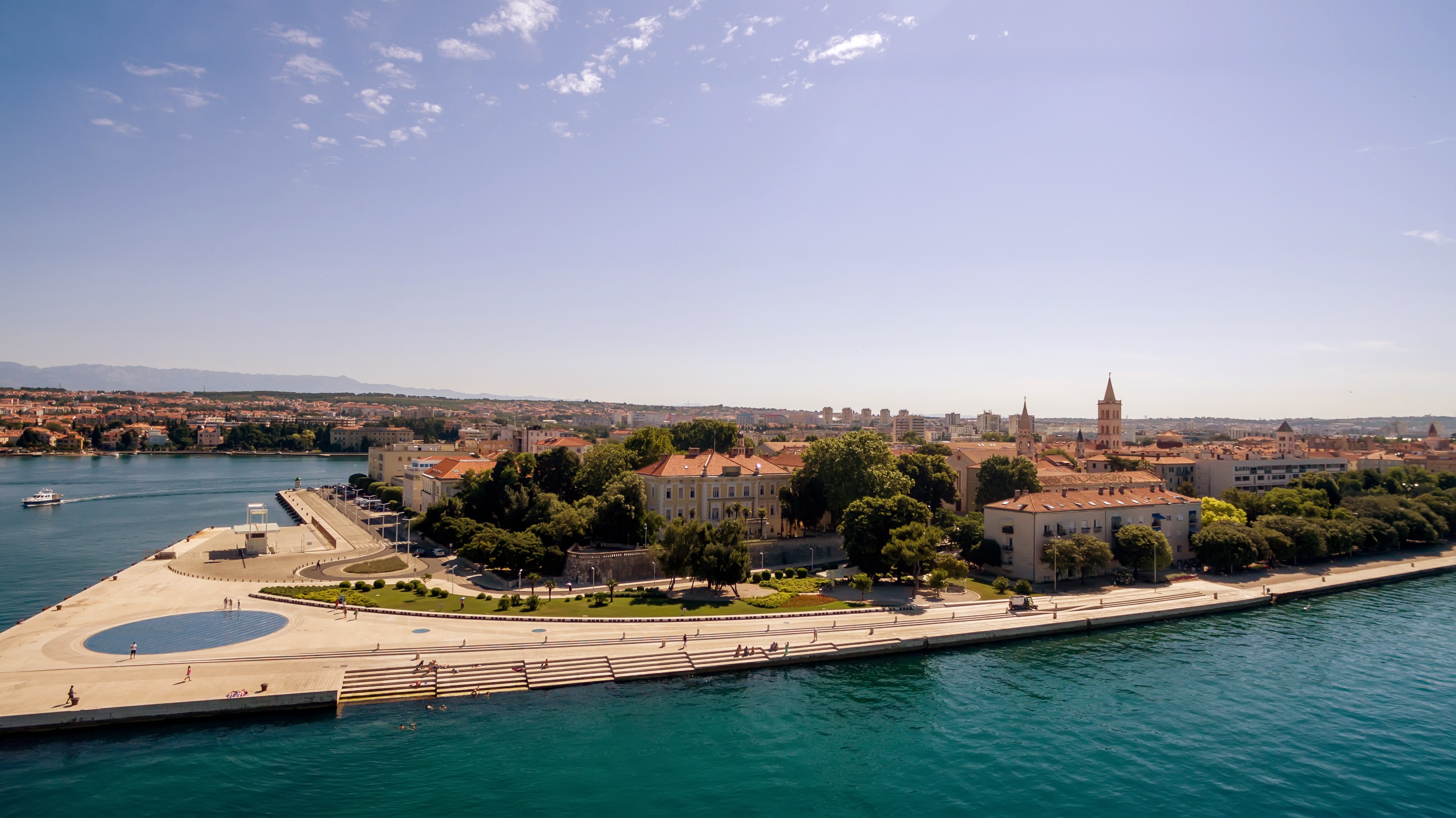 Panoramski zračni pogled na Zadar u Hrvatskoj, prikazujući rivu s poznatim Morskim orguljama i instalacijom Pozdrav Suncu, tirkizno Jadransko more i siluetu povijesnog starog grada pod vedrim plavim nebom.