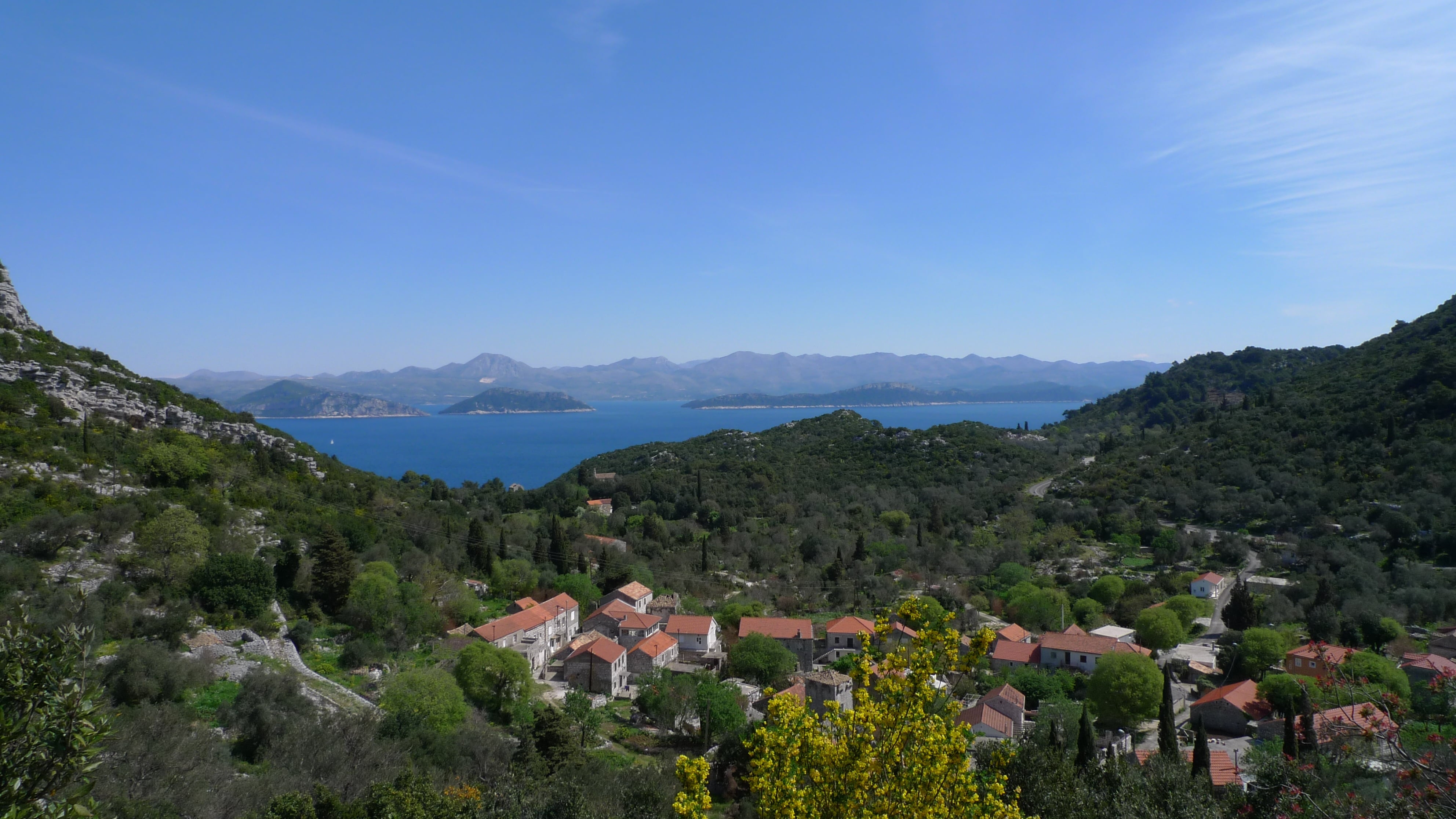 Panoramic view of a traditional village surrounded by lush green hills on Mljet Island, overlooking the blue Adriatic Sea, nearby islands, and distant mountains.