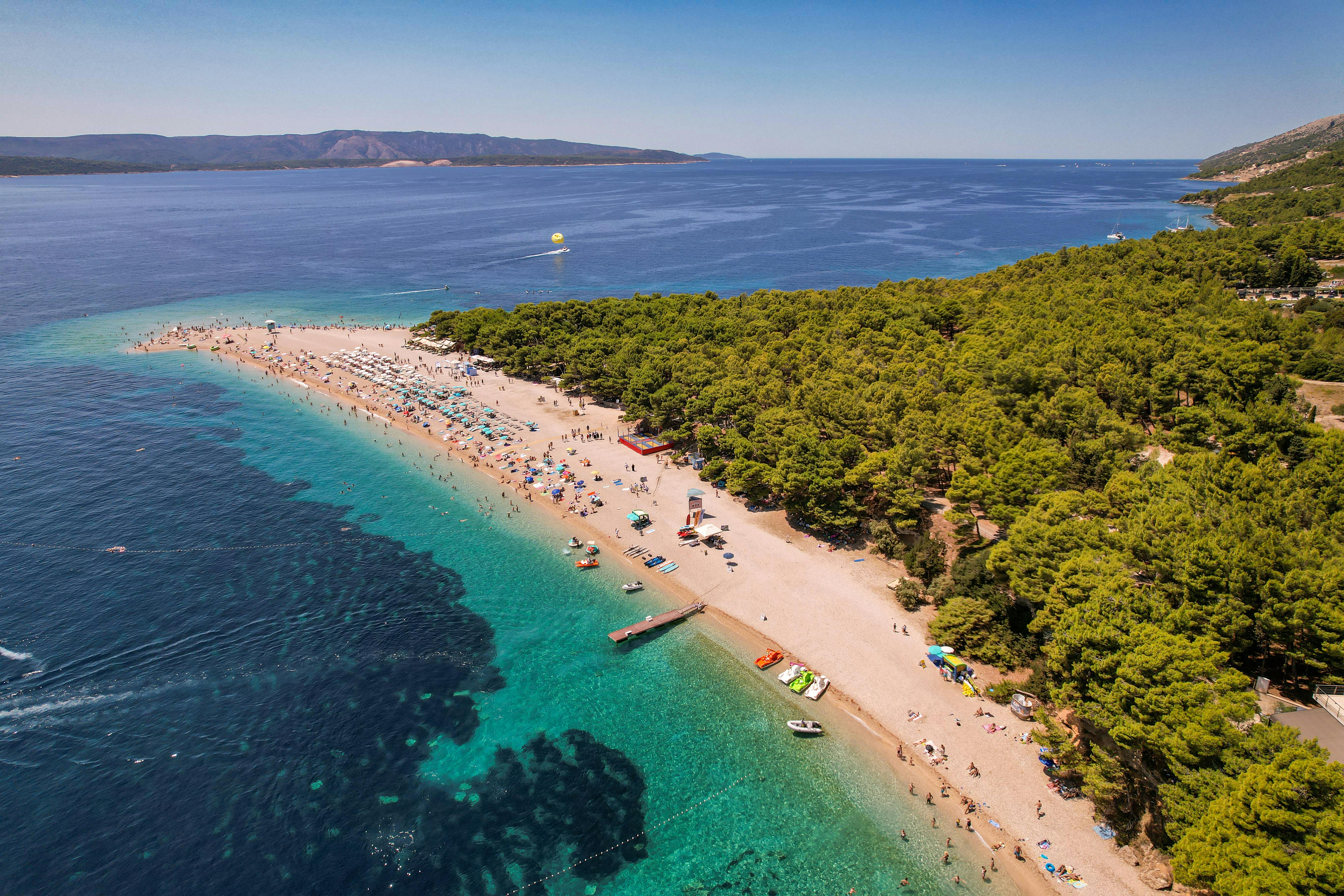Luftfoto af den ikoniske Zlatni Rat-strand på øen Brač, Kroatien, der strækker sig ud i det krystalklare Adriaterhav. Den gyldne pebble-kystlinje er omgivet af frodige pinjeskove og turkist vand, med besøgende, der nyder svømning, solbadning og vandsport mod en betagende kystbaggrund.