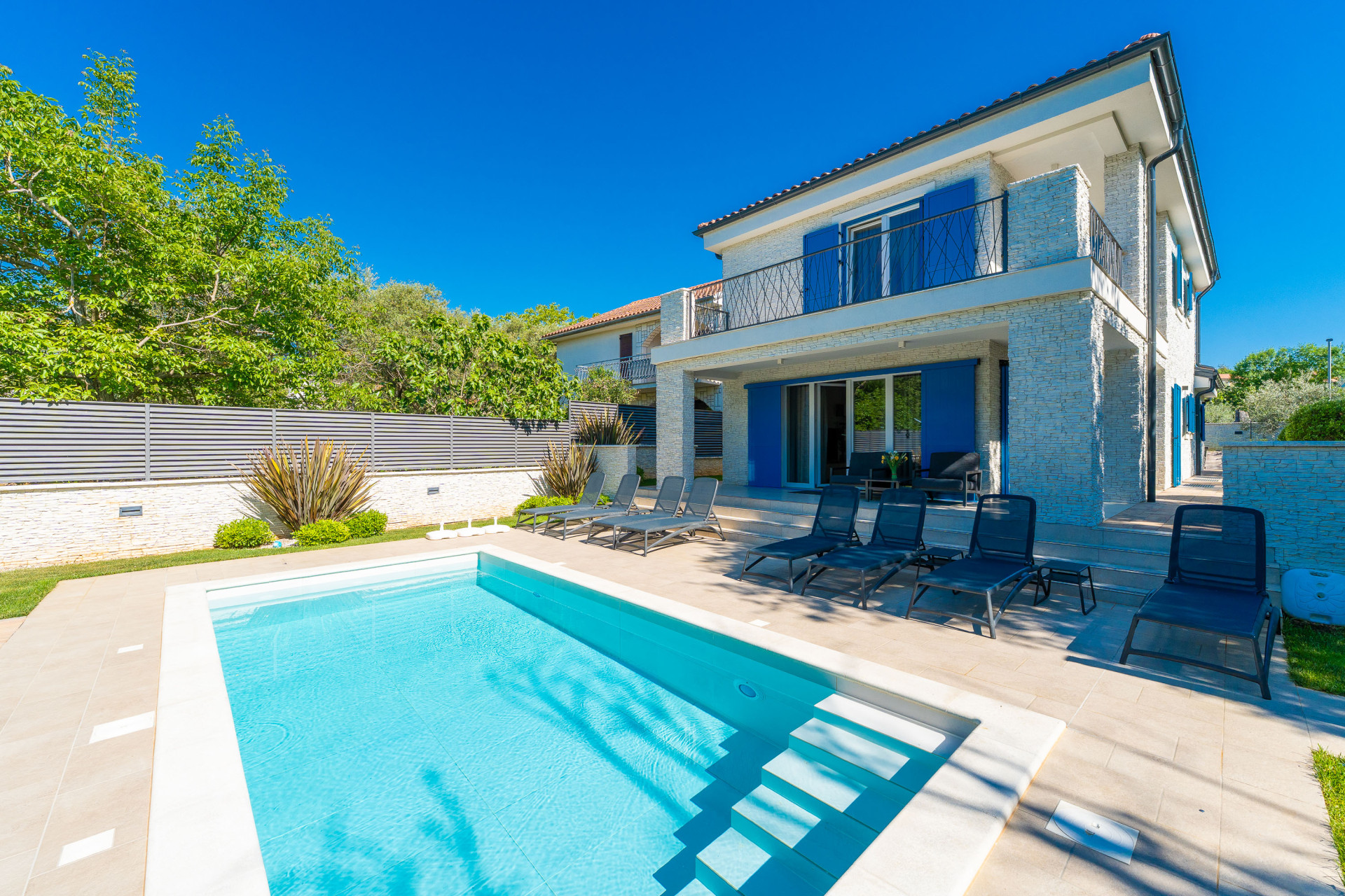 Moderna Villa Valle sull'isola di Krk con piscina privata, elegante architettura mediterranea, caratteristiche persiane blu, lettini prendisole e spaziosa terrazza esterna sotto un cielo limpido.
