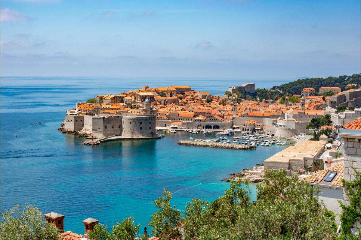 Veduta panoramica della Città Vecchia di Dubrovnik con mura medievali in pietra, mare Adriatico turchese e tetti storici arancioni sulla costa dalmata.