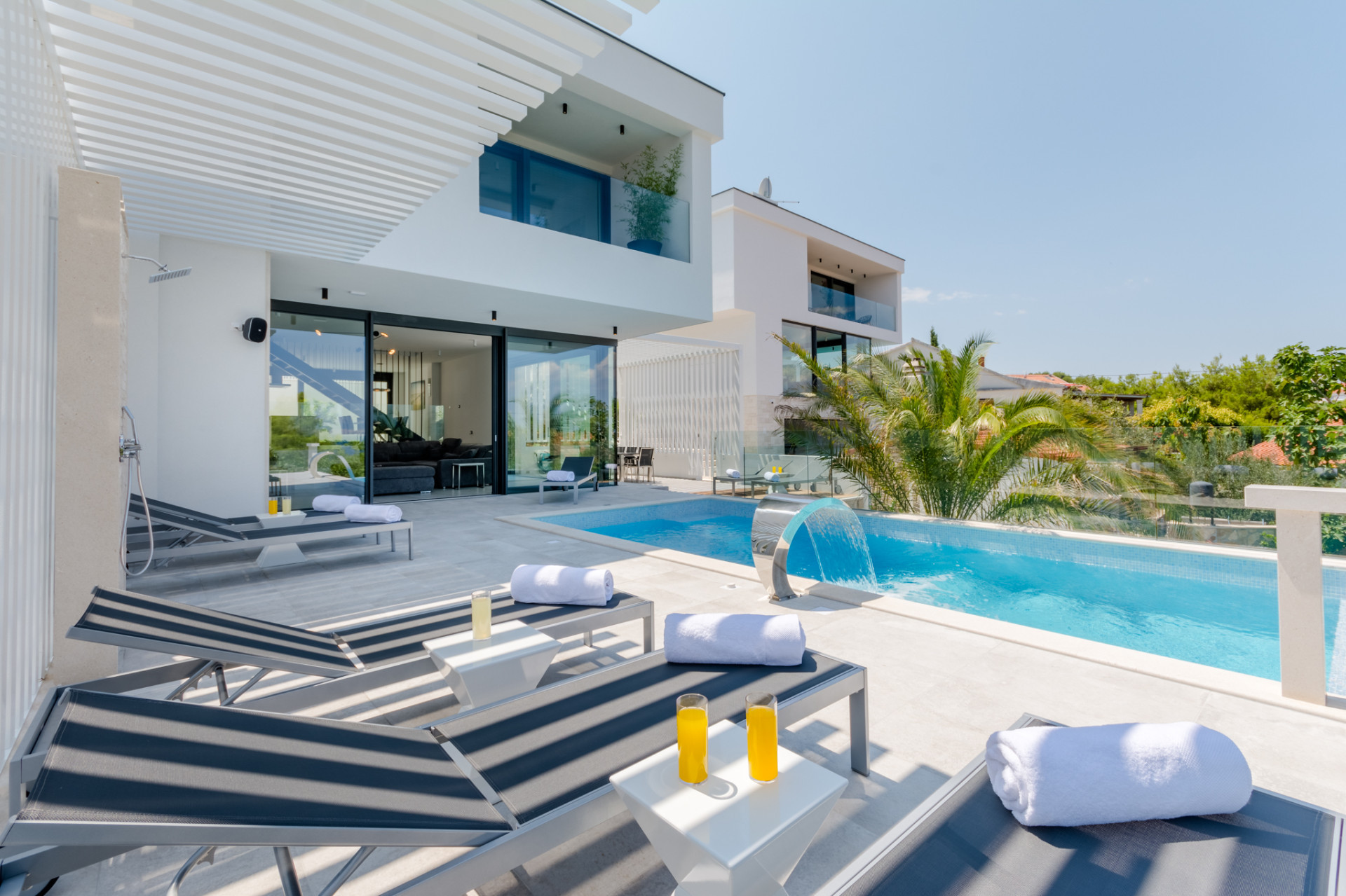 Villa Pax on Brač Island, featuring a private swimming pool with a waterfall feature, modern architecture, a spacious sun terrace with loungers and a shaded outdoor relaxation area.