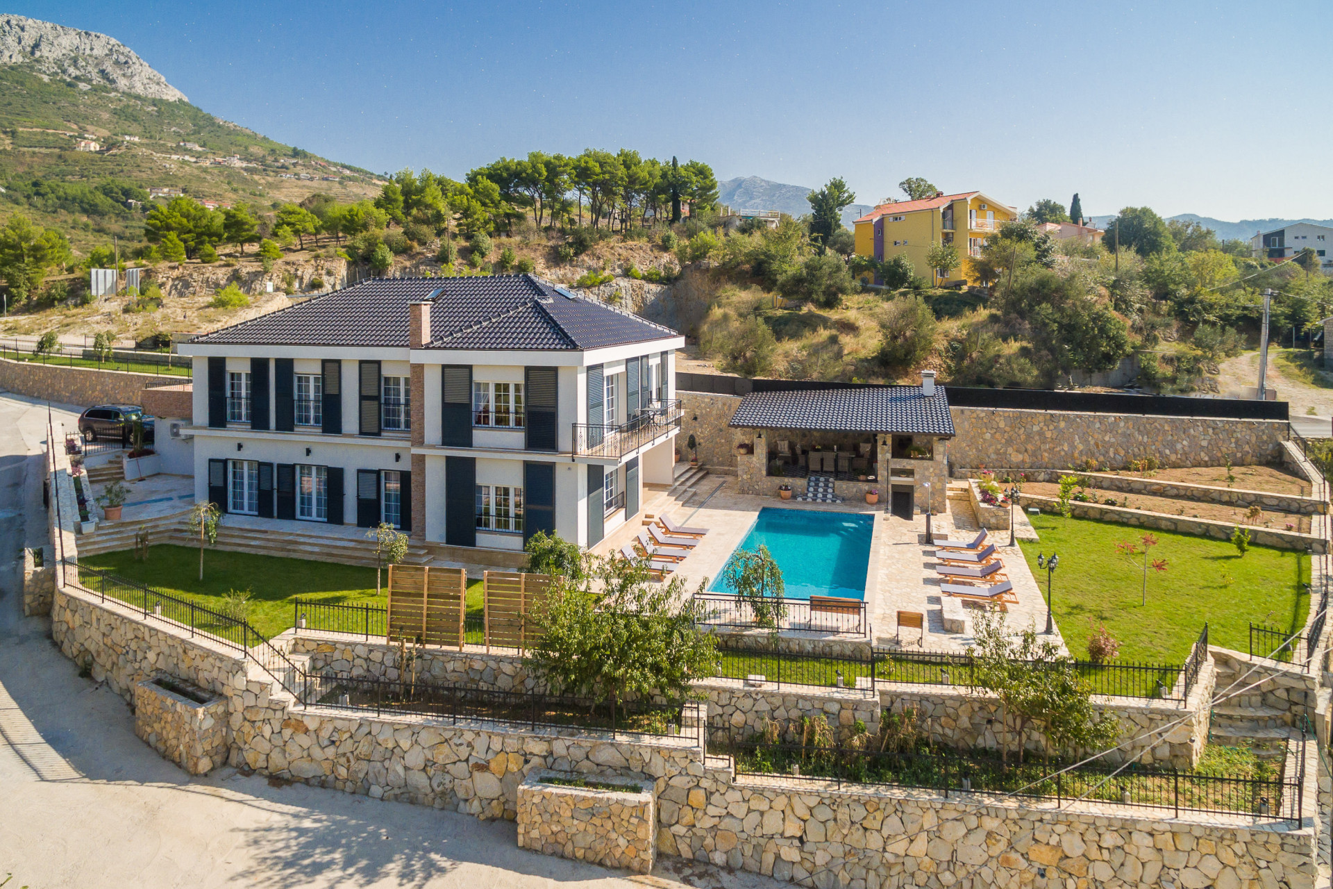 Aerial view of Villa Zvonimir, featuring a private swimming pool, landscaped garden, sun loungers, and a spacious stone terrace surrounded by a scenic mountain landscape.