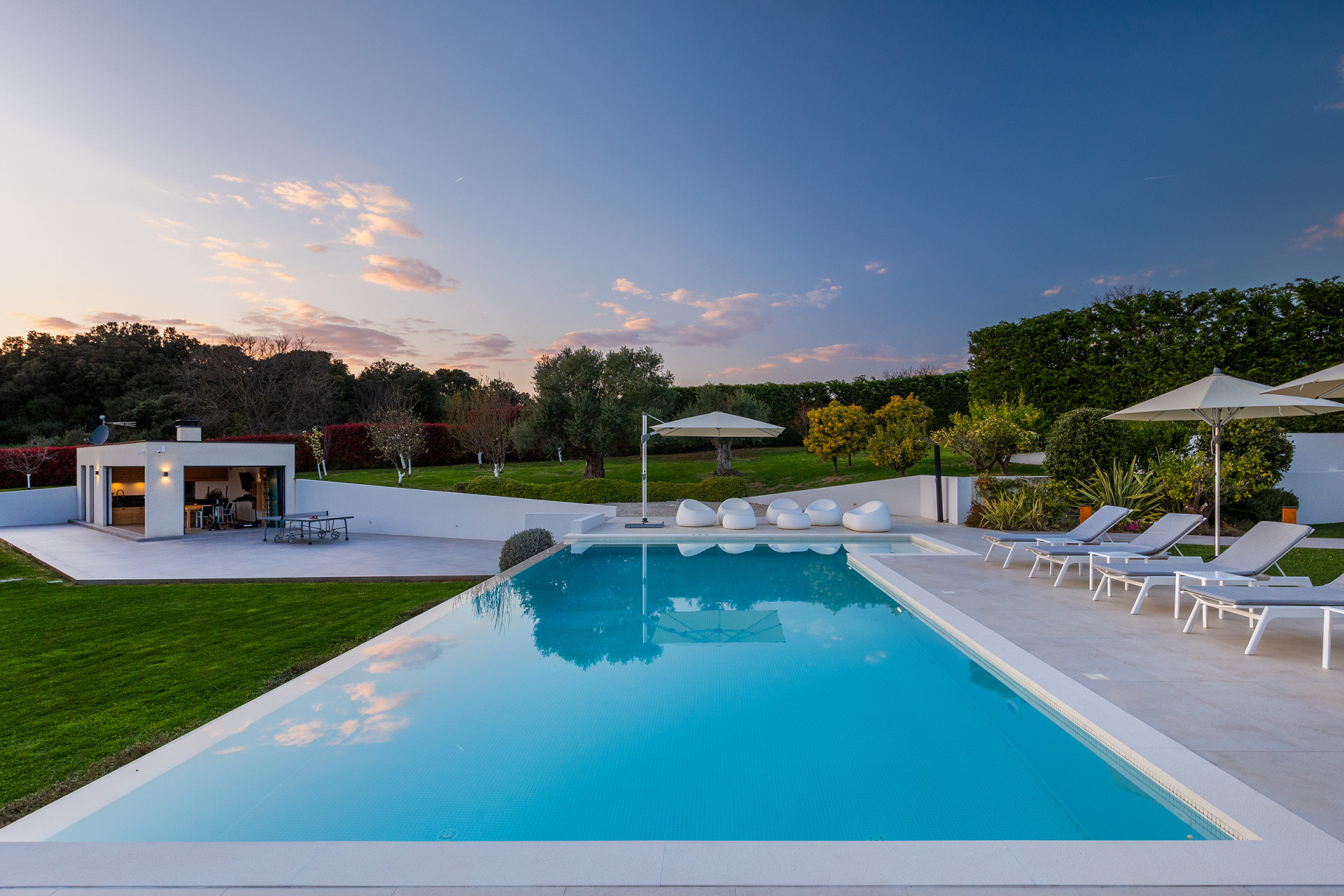 Twilight view of Villa PS Deluxe, a modern luxury villa in Croatia featuring a large illuminated infinity pool, elegant sun terrace with loungers and umbrellas, beautifully landscaped garden, and stylish outdoor lounge area at sunset.
