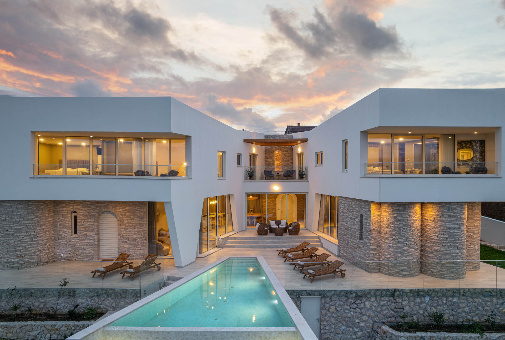 Twilight view of Villa Di Polisane, a contemporary luxury villa in Croatia featuring a long illuminated private swimming pool, spacious sun terrace with wooden loungers, floor-to-ceiling glass walls, elegant stone architecture, and warm ambient lighting beneath a dramatic sunset sky.