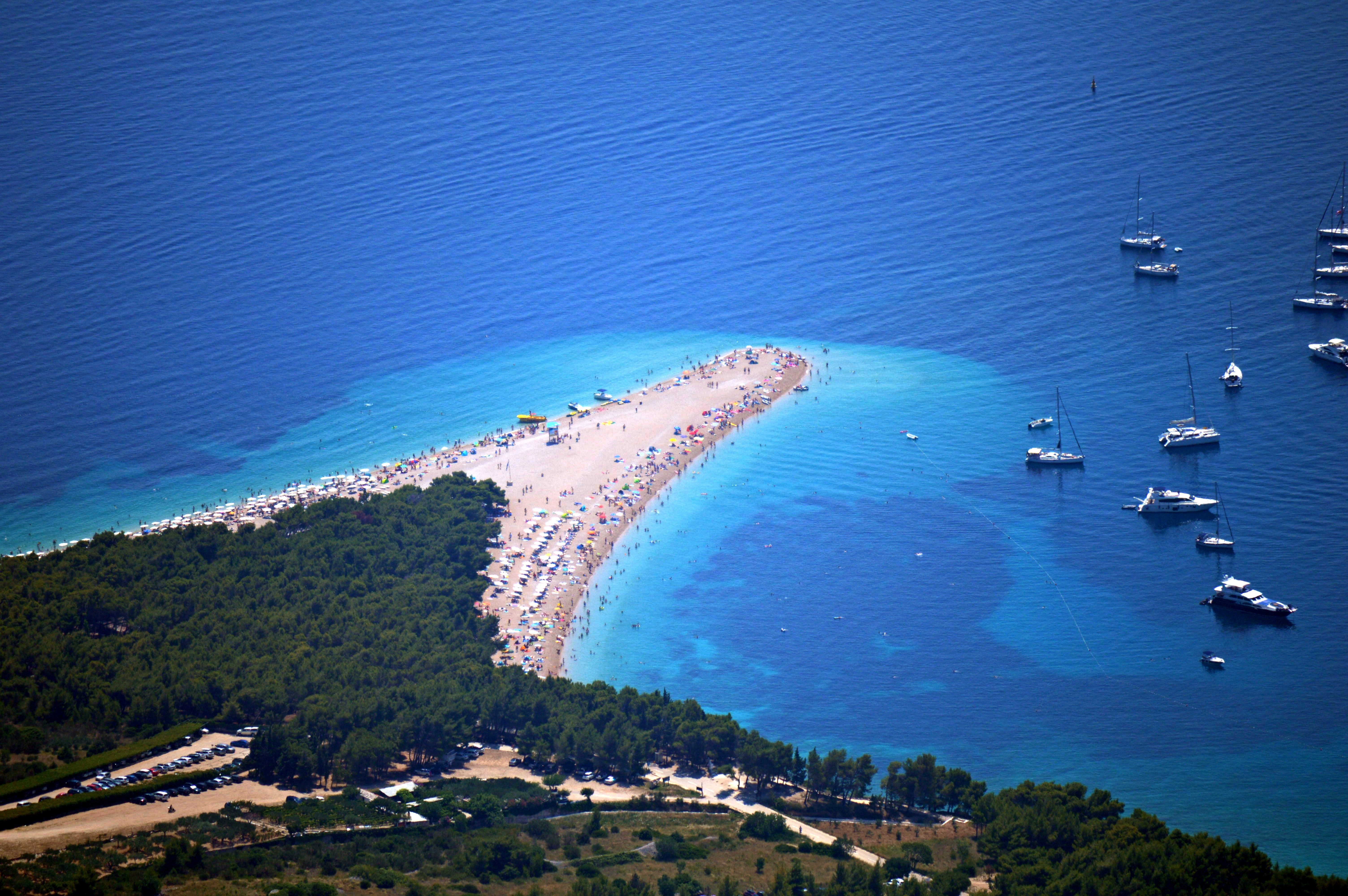Aerial view of Zlatni Rat Beach on Brač Island, Croatia, featuring its distinctive pebble shoreline, turquoise Adriatic Sea, surrounding pine forest and yachts anchored offshore.