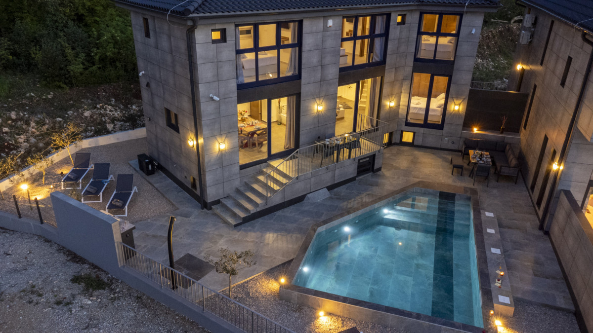 Vista dall'alto di Villa Signature Krk al crepuscolo, con architettura in pietra, piscina riscaldata e pareti vetrate che rivelano l'elegante interno.