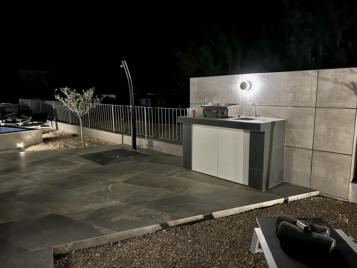 Moderna cucina esterna presso Villa Signature Krk di notte, con un barbecue a gas plancha professionale, un lavandino e un'elegante doccia esterna.