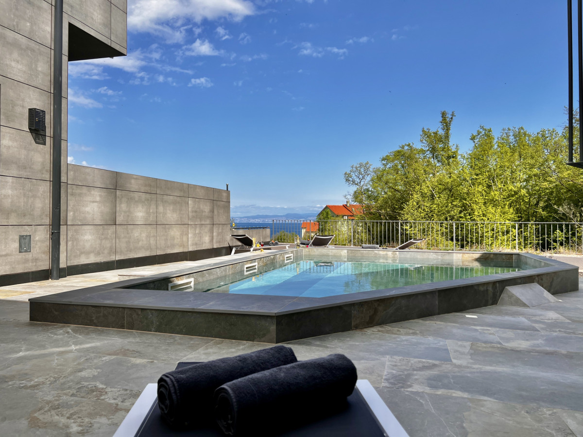 Rilassante terrazza presso Villa Signature Krk, con un'elegante piscina con bordo scuro, lussuosi asciugamani arrotolati e vista sul mare in lontananza sotto un cielo blu brillante.