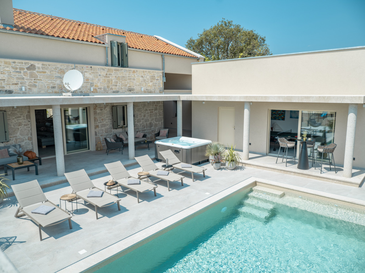 Exterior view of Villa Utopia featuring a private turquoise pool, a sun deck with loungers, a jacuzzi, a modern slatted pergola, and the elegant stone facade.