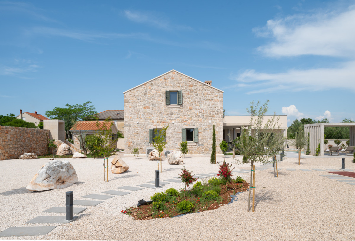 Entrance view of Villa Utopia, featuring a rustic stone exterior, a manicured gravel courtyard with stepping stones, decorative olive trees, and modern lighting fixtures, all set against a clear blue sky.