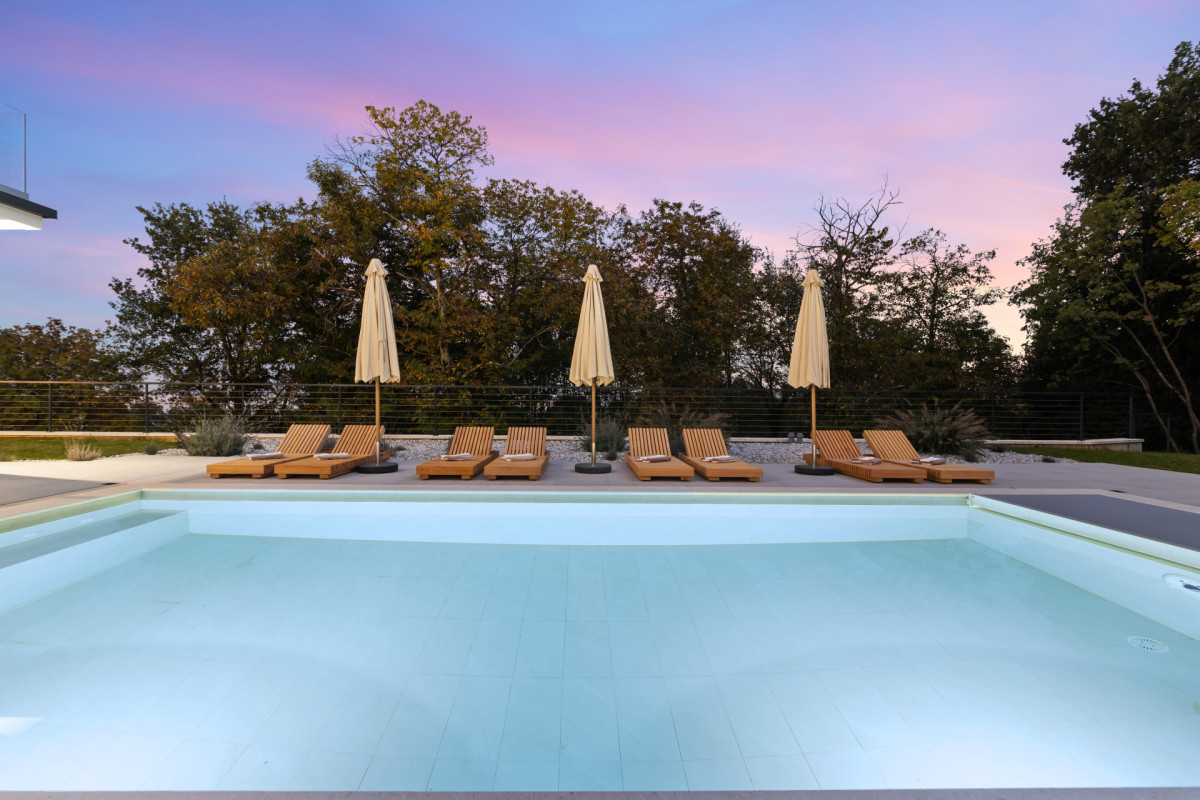 Entspannungsbereich der Villa Casa Calme: Hochwertige Holz-Sonnenliegen und Sonnenschirme am kristallklaren Pool während eines rosafarbenen Sonnenuntergangs.