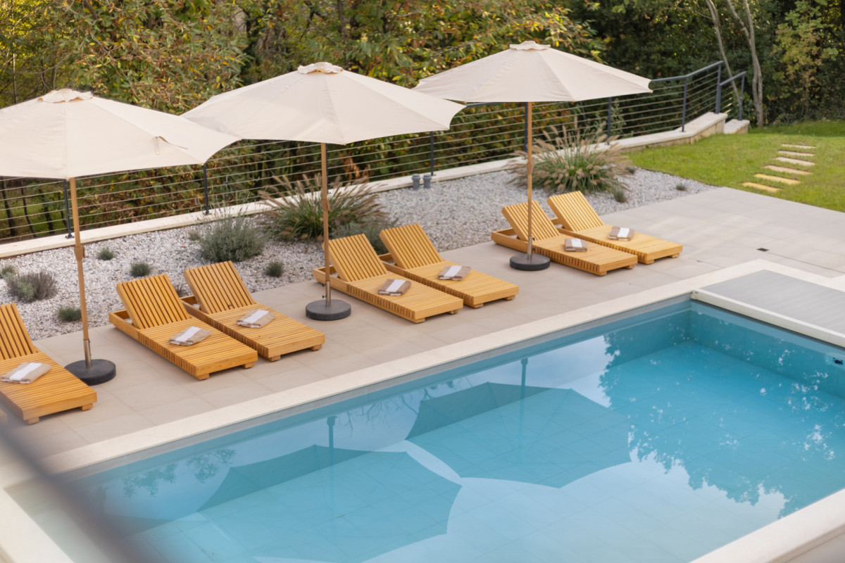 Luxuriöses Pooldeck in der Villa Casa Calme mit hochwertigen Sonnenliegen aus Holz, frischen Handtüchern, beige farbenen Sonnenschirmen und einem kristallklaren Swimmingpool.