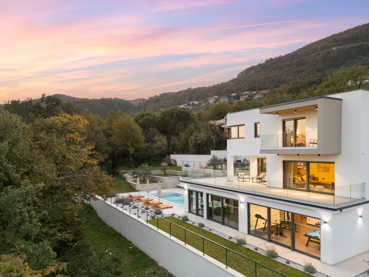 Seitenansicht der Villa Casa Calme bei Sonnenuntergang, die die mehrstöckige moderne Architektur, den Außenpool und die großen Glasfronten zeigt, die den Blick auf den hauseigenen Fitnessraum und den Loungebereich freigeben.