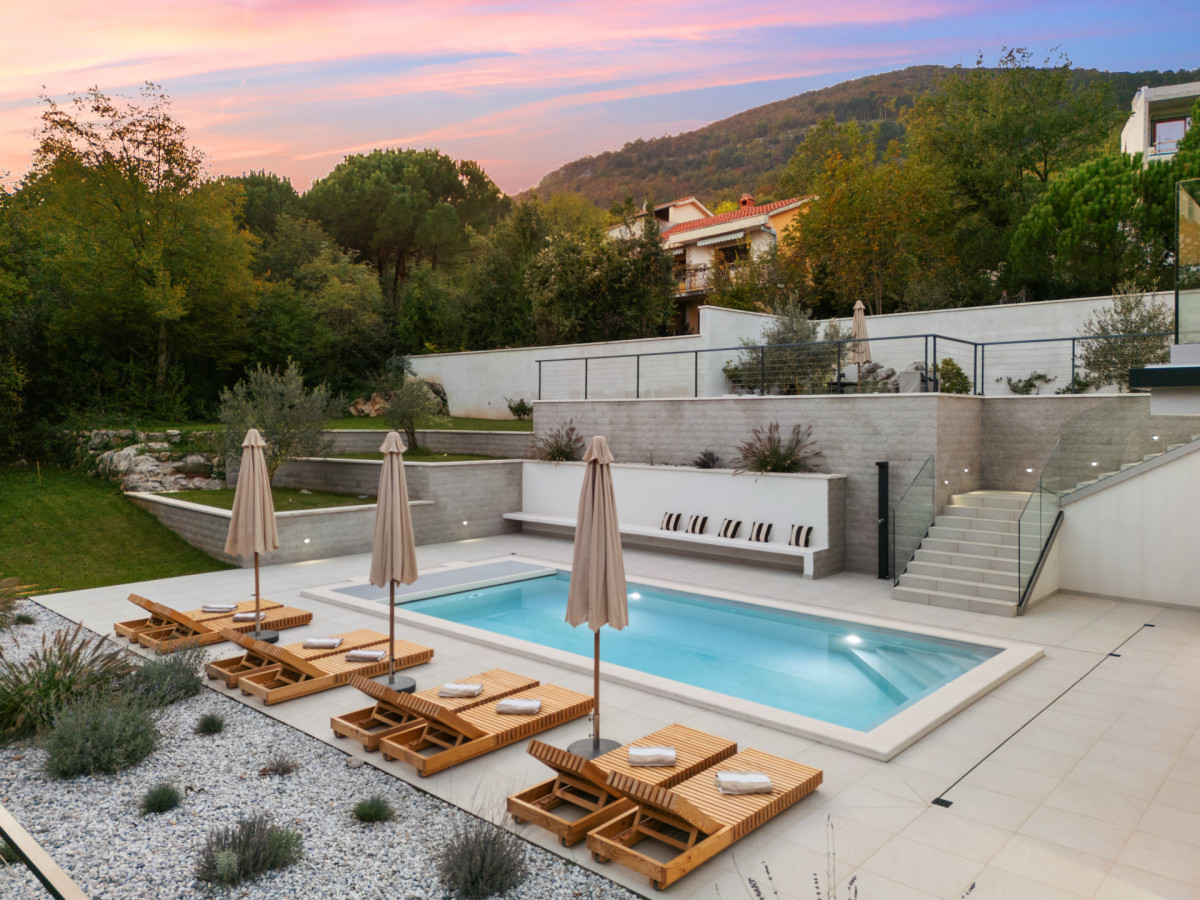 Abendstimmung in der Villa Casa Calme: ein beleuchteter Poolbereich und ein elegantes Sonnendeck unter einem wunderschönen rosa-orangen Sonnenuntergang, die die friedliche Umgebung der Villa unterstreichen.
