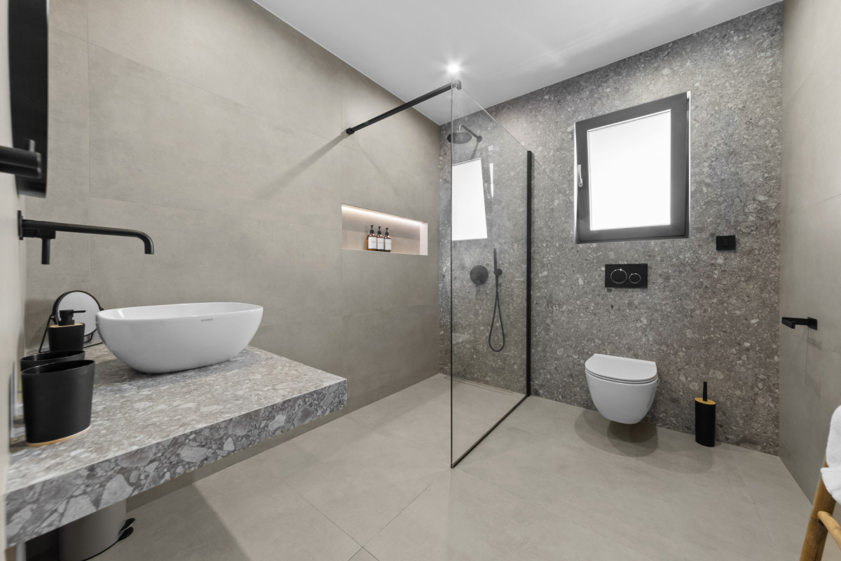 Modernes Luxusbadezimmer in der Villa Casa Calme mit begehbarer Dusche und Glastrennwand, grauen Fliesen in Steinoptik, einem Aufsatzwaschbecken und eleganten schwarzen Armaturen.
