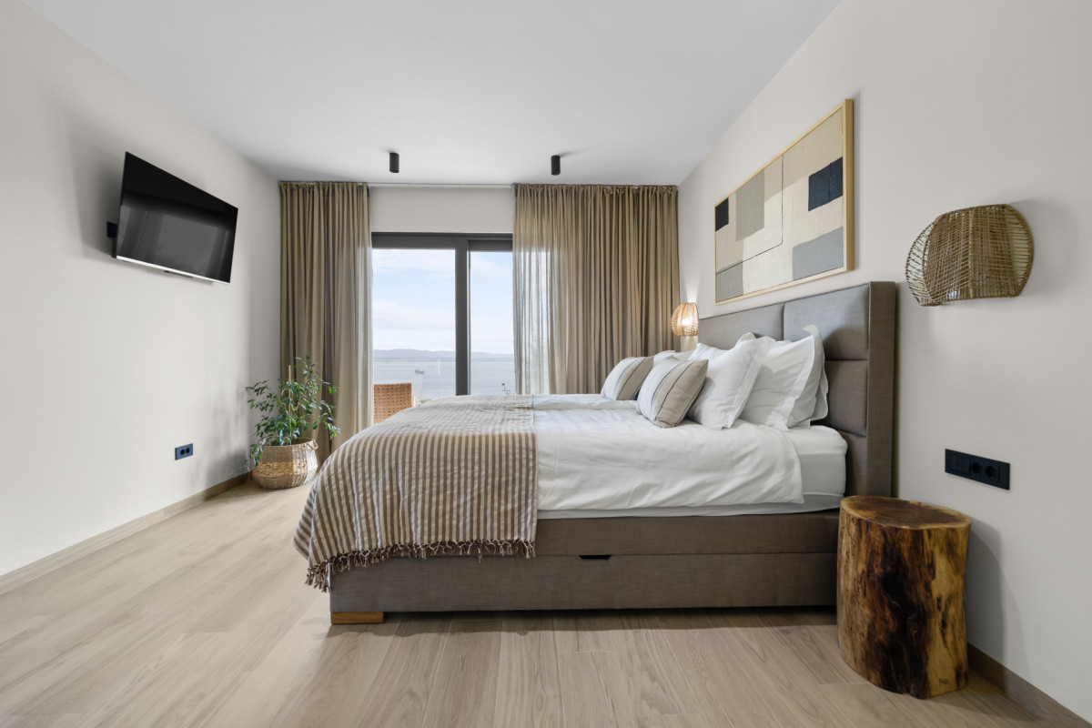 Helles Schlafzimmer mit eigenem Bad in der Villa Casa Calme: mit einem großen Bett mit hochwertiger Bettwäsche, einem wandmontierten Fernseher und raumhohen Glastüren, die auf einen Balkon mit malerischem Meerblick führen.