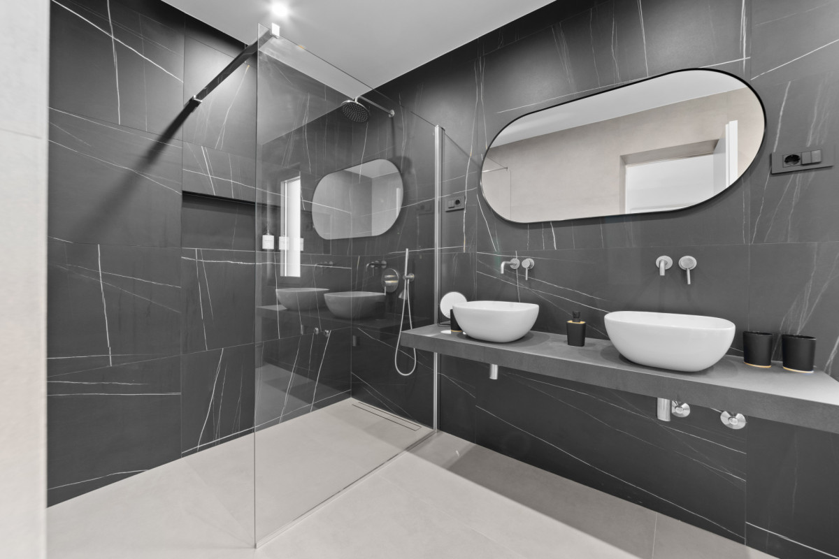 Modernes En-Suite-Badezimmer mit einem „His and Hers“-Doppelwaschbecken, einem eleganten ovalen Spiegel und schlichten, minimalistischen Armaturen vor dunklen Premium-Fliesen u der Villa Casa Calme.