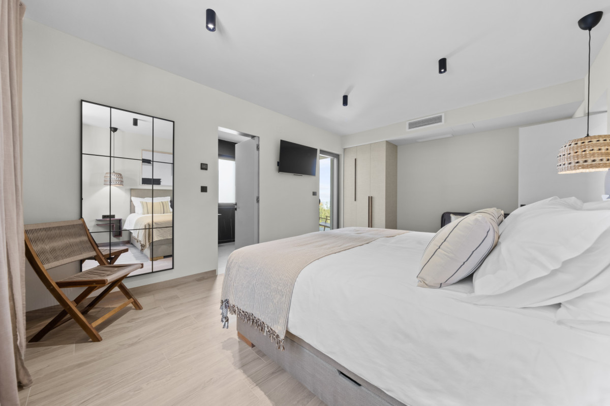 Großzügiges Schlafzimmer mit eigenem Bad in der Villa Casa Calme, ausgestattet mit einem großen, bequemen Bett, einem stilvollen Sprossenspiegel, einem Lounge-Sessel aus Holz und einem Blick in das elegante private Badezimmer.