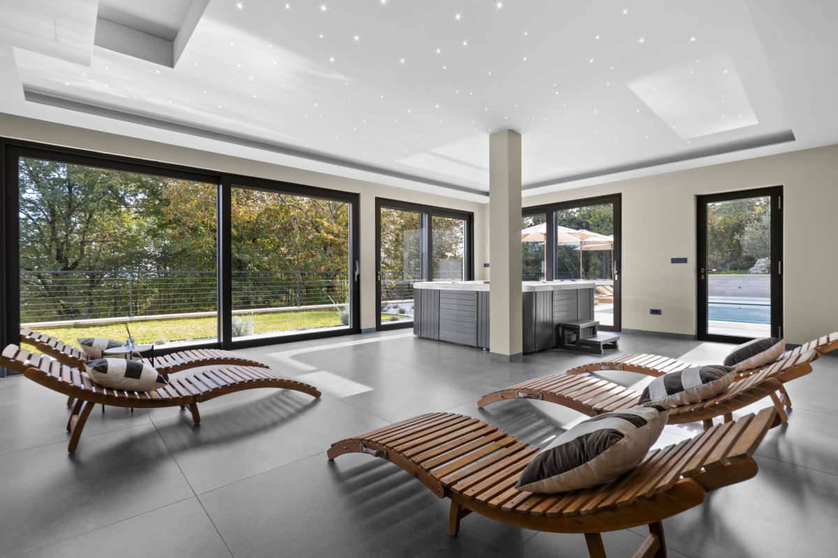 Ruhige Indoor-Entspannungszone in der Villa Casa Calme: stilvolle Holzliegen und ein Whirlpool-Bereich, umgeben von Panoramafenstern mit Blick auf das mediterrane Grün.